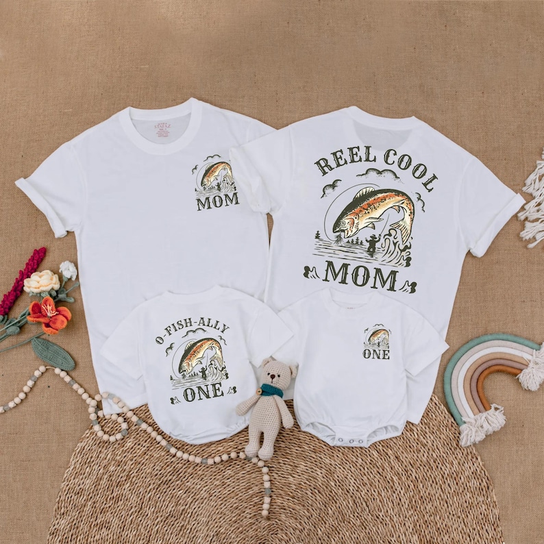 May include: White t-shirts and onesies with fishing-themed graphics. One shirt says "Reel Cool Mom" with a fish jumping out of the water. Another says "Mom" with a fish graphic. The onesies say "One" and "O-Fish-Ally One".