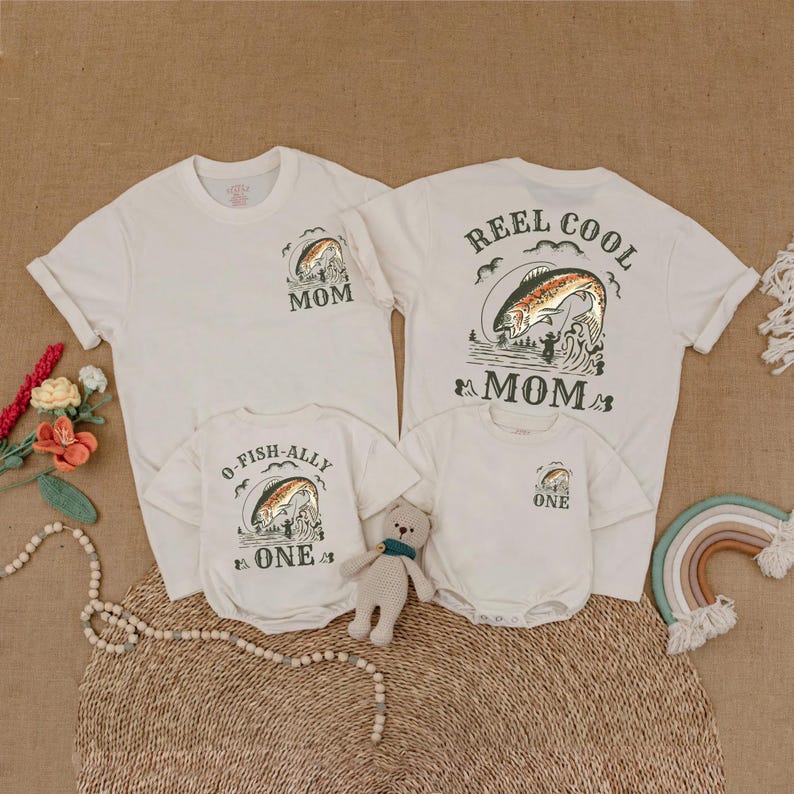 May include: Off-white t-shirts and onesies with a fishing-themed graphic. The shirts say "Mom" and "Reel Cool Mom" on the front and back, and the onesies say "O-Fish-Ally One" and "One". A family set of matching apparel.