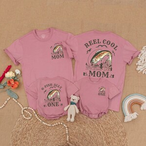 May include: Pink family matching t-shirts and a onesie with fishing-themed graphics. The shirts say "Mom" and "Reel Cool Mom" with a fish jumping out of the water. The onesie and a shirt say "O-Fish-Ally One" and "One".
