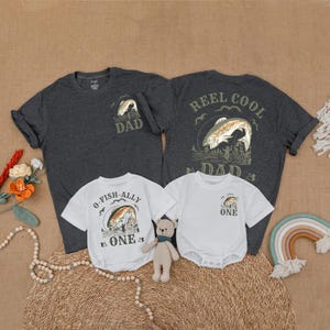May include: A collection of matching fishing-themed shirts and onesies. Two dark gray t-shirts with "DAD" and "REEL COOL DAD" graphics. Two white onesies and a t-shirt with "O-FISH-ALLY ONE" and "ONE" graphics. A neutral-toned teddy bear and rainbow decor are also present.