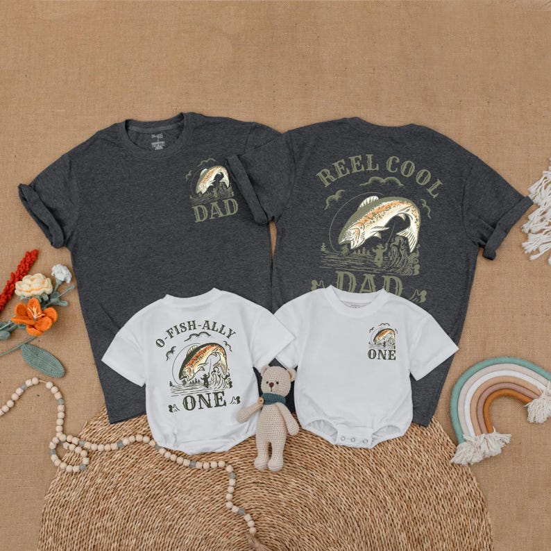 May include: A collection of matching fishing-themed shirts and onesies. Two dark gray t-shirts with "DAD" and "REEL COOL DAD" graphics. Two white onesies and a t-shirt with "O-FISH-ALLY ONE" and "ONE" graphics. A neutral-toned teddy bear and rainbow decor are also present.