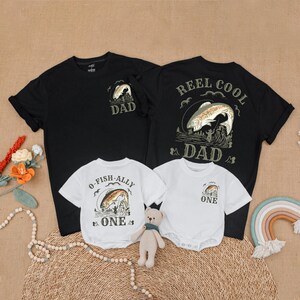 May include: Black and white t-shirts and white onesies with fishing-themed graphics. The adult shirts say "DAD" and "REEL COOL DAD." The onesies say "O-FISH-ALLY ONE" and "ONE." A small teddy bear sits in front of the shirts.