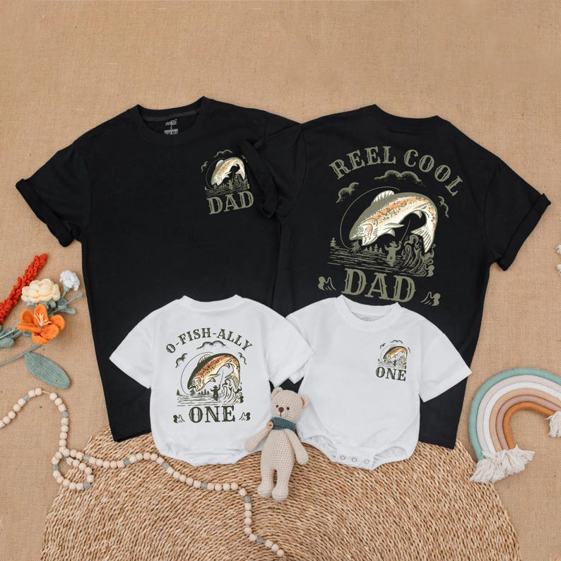 May include: Black and white t-shirts and white onesies with fishing-themed graphics. The adult shirts say "DAD" and "REEL COOL DAD." The onesies say "O-FISH-ALLY ONE" and "ONE." A small teddy bear sits in front of the shirts.