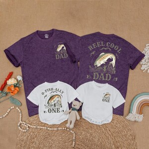 May include: Purple and white matching t-shirts and onesies with fishing-themed graphics. The adult shirts say "Dad" and "Reel Cool Dad." The baby onesies say "O-Fish-Ally One" and "One." A small teddy bear and a rainbow decoration are also in the image.