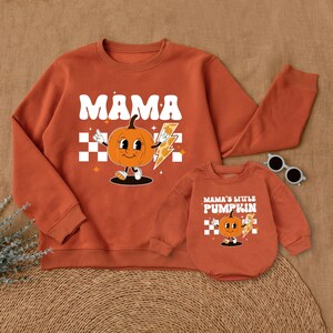 May include: Orange and white matching sweatshirts with a pumpkin graphic. The adult sweatshirt says "Mama" and the child sweatshirt says "Mama's Little Pumpkin".