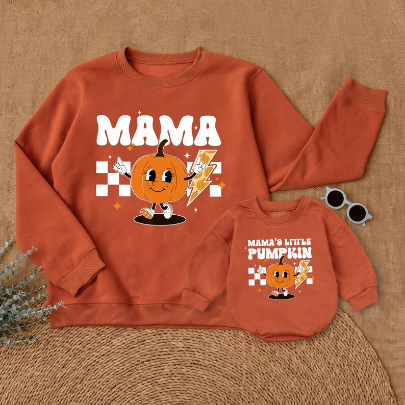May include: Orange and white matching sweatshirts with a pumpkin graphic. The adult sweatshirt says "Mama" and the child sweatshirt says "Mama's Little Pumpkin".