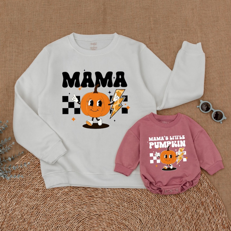 May include: A white sweatshirt with the text "MAMA" and a cartoon pumpkin with a lightning bolt. A pink long-sleeve onesie with the text "MAMA'S LITTLE PUMPKIN" and a cartoon pumpkin with a lightning bolt.