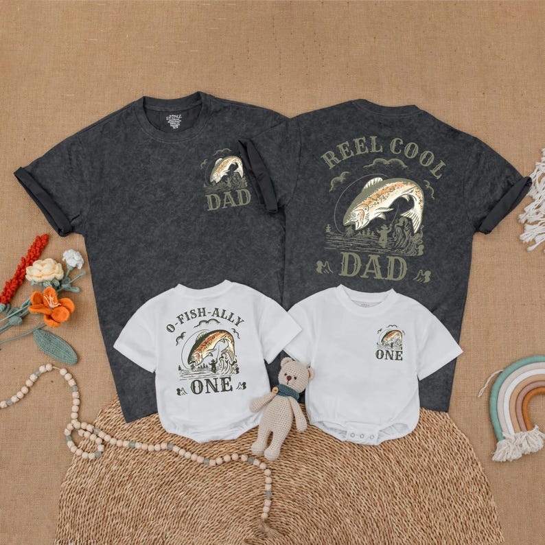 May include: A family matching set of fishing-themed apparel. Includes a dark gray t-shirt with "DAD" and a fish graphic, a white onesie with "O-FISH-ALLY ONE", and a white onesie with "ONE". A teddy bear and rainbow decor are also present.
