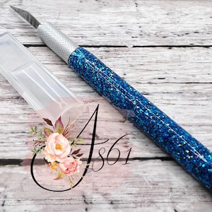 May include: A blue glitter craft knife with a silver blade and a clear plastic case. The knife has a blue glitter finish with iridescent sparkles.