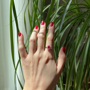 May include: A hand with red nail polish is wearing a ring with a red gemstone and small diamonds. The hand is in front of a green plant.