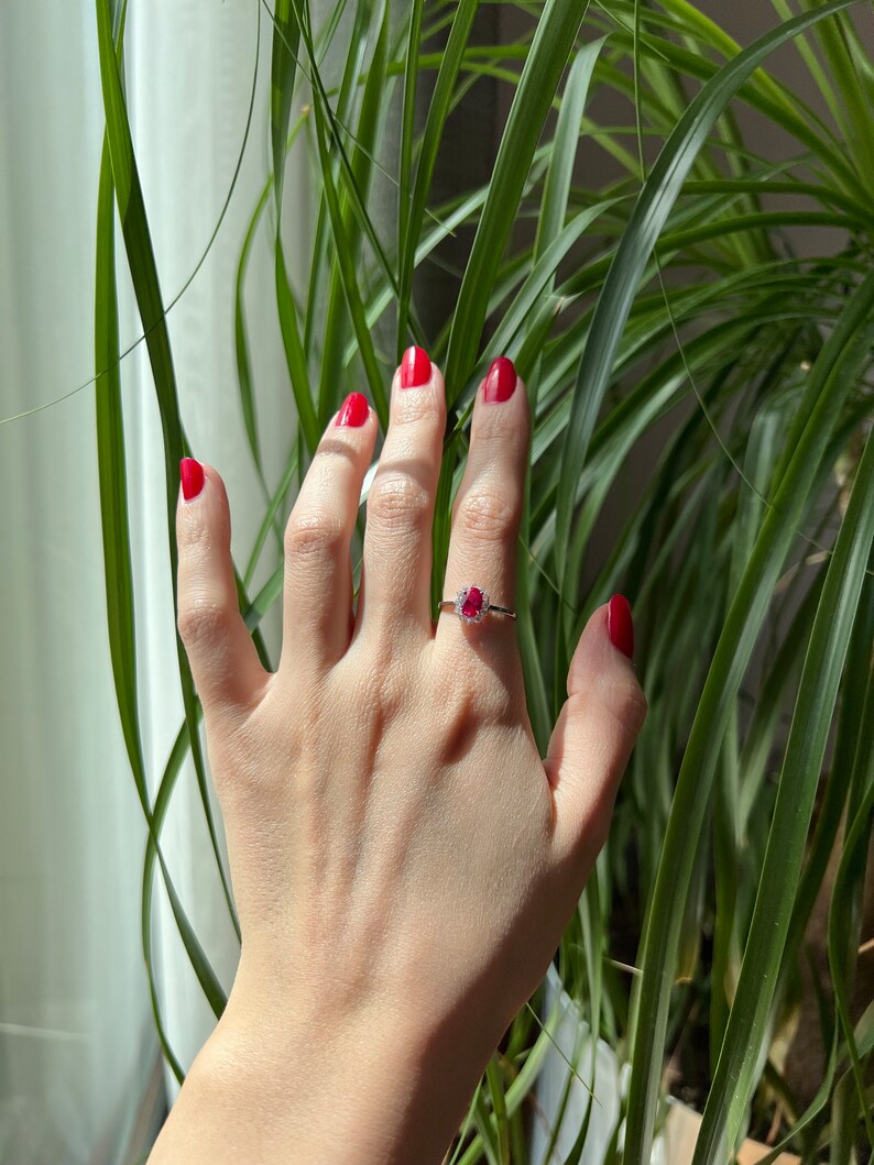 May include: A hand with red nail polish is wearing a ring with a red gemstone and small diamonds. The hand is in front of a green plant.