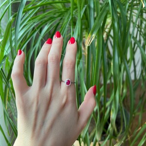 May include: A close-up of a hand wearing a ring with a red gemstone surrounded by smaller white gemstones. The hand has red nail polish and is resting against a green plant.
