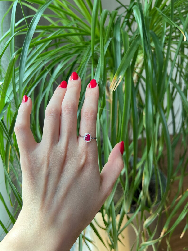 May include: A close-up of a hand wearing a ring with a red gemstone surrounded by smaller white gemstones. The hand has red nail polish and is resting against a green plant.
