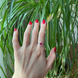May include: A silver ring with a red gemstone surrounded by smaller white gemstones. The ring is on a person's finger.