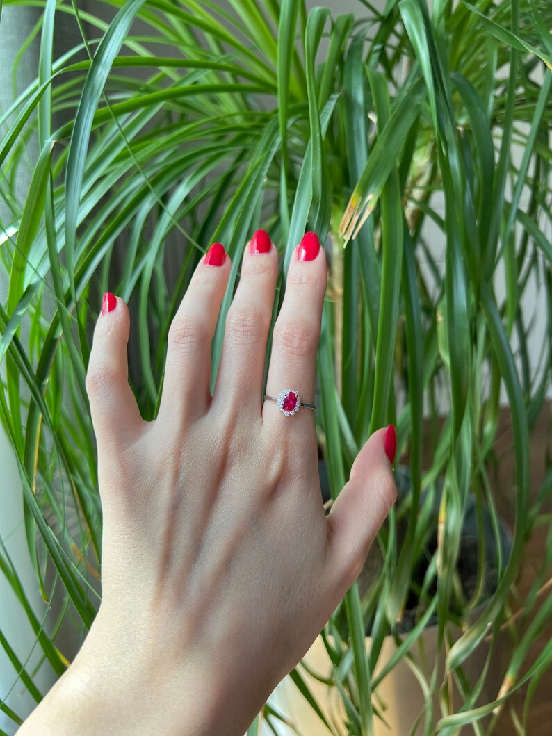 May include: A silver ring with a red gemstone surrounded by smaller white gemstones. The ring is on a person's finger.