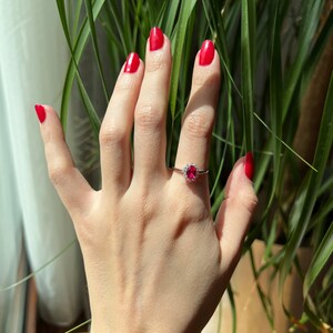 May include: A hand with red nail polish is wearing a ring with a large red gemstone surrounded by smaller white gemstones.