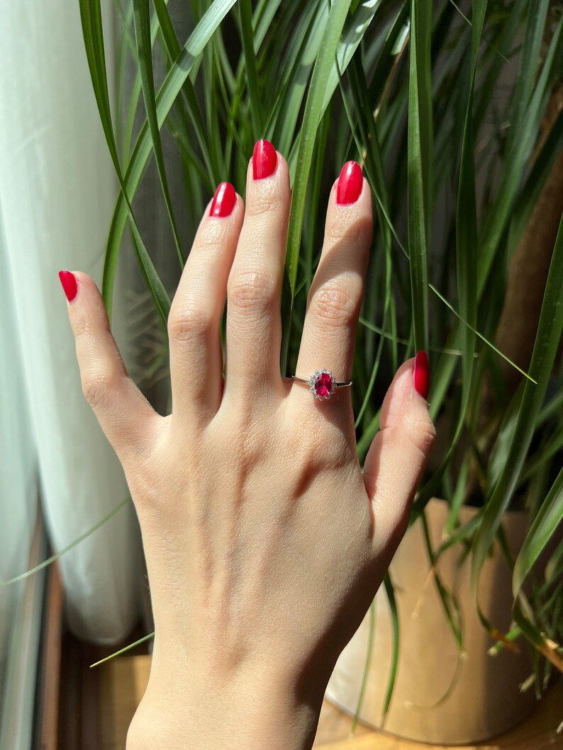 May include: A hand with red nail polish is wearing a ring with a large red gemstone surrounded by smaller white gemstones.