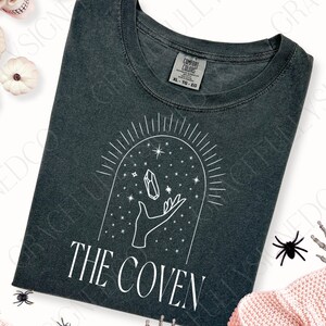 May include: A dark gray t-shirt with a white graphic design of a hand holding a crystal under a starry sky with the text "THE COVEN".