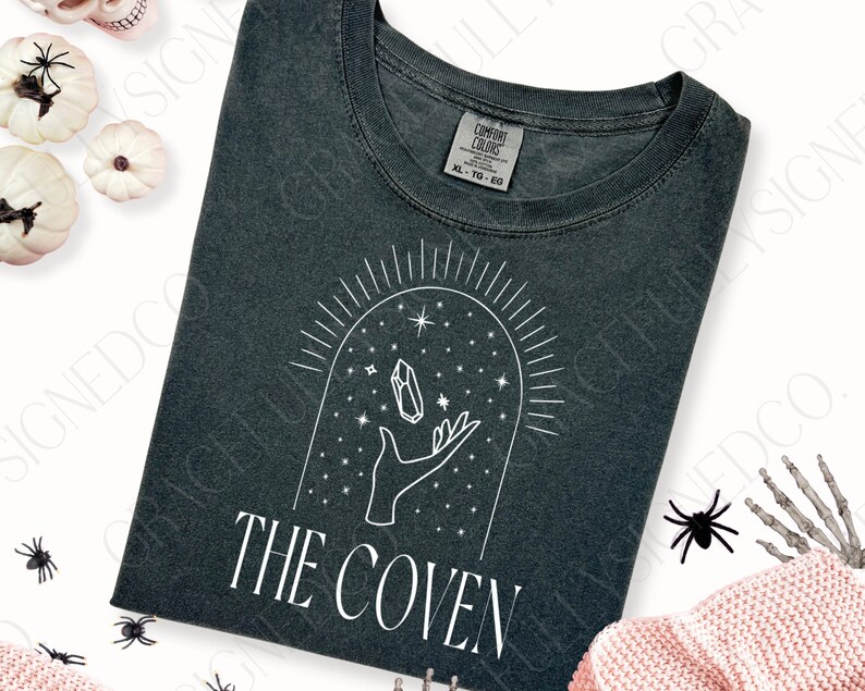 May include: A dark gray t-shirt with a white graphic design of a hand holding a crystal under a starry sky with the text "THE COVEN".
