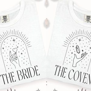 May include: Two white t-shirts with black line drawings of hands holding a heart and a crystal. The text "The Bride" and "The Coven" are printed on the shirts.