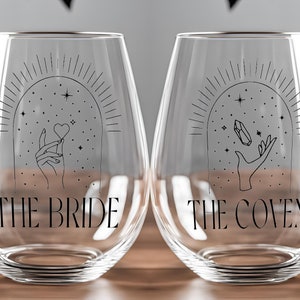 May include: Two clear glass wine glasses with black text and illustrations. The glass on the left says "The Bride" and features a hand holding a heart. The glass on the right says "The Coven" and features a hand holding a crystal.