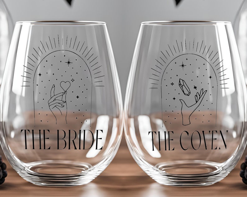 May include: Two clear glass wine glasses with black text and illustrations. The glass on the left says "The Bride" and features a hand holding a heart. The glass on the right says "The Coven" and features a hand holding a crystal.