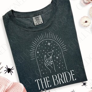 May include: A dark gray t-shirt with a white graphic design of a hand holding a heart under a starry arch. The text "THE BRIDE" is printed below the graphic.
