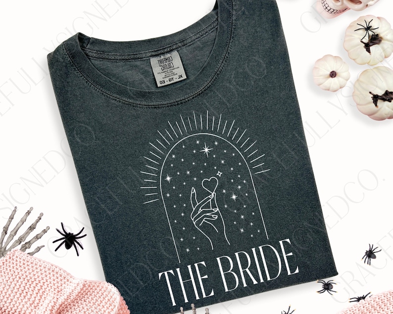 May include: A dark gray t-shirt with a white graphic design of a hand holding a heart under a starry arch. The text "THE BRIDE" is printed below the graphic.