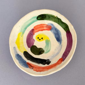 Op de afbeelding: Een klein wit keramiek schaaltje met een kleurrijk swirl-ontwerp. Het ontwerp heeft gele, groene, blauwe, rode, paarse en zwarte verf. Een klein geel smiley-gezicht bevindt zich in het midden van de swirl.