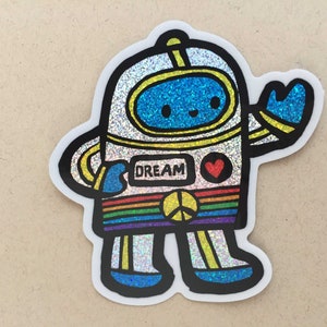 Pode incluir: Um adesivo de astronauta branco coberto de glitter com uma faixa arco-íris, um símbolo de paz e a palavra "DREAM" no peito. O astronauta tem o rosto azul e está acenando.