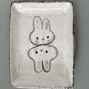 May include: A rectangular ceramic tray with a white speckled glaze and a dark brown textured rim. The center features a hand-painted illustration of two stacked grey rabbits, one upright and one inverted, with simple black facial features.