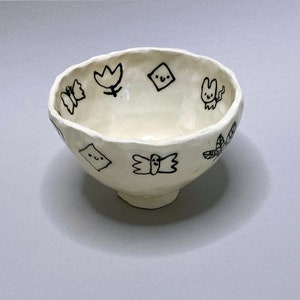 May include: A white ceramic bowl with a textured surface and black hand-drawn designs of butterflies, flowers, and other whimsical shapes.
