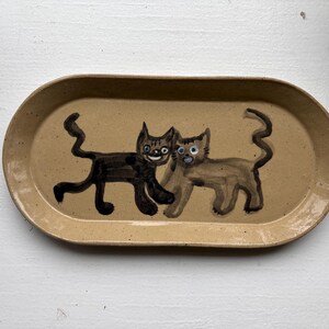 May include: An oval-shaped ceramic tray with a speckled tan glaze. The center features a hand-painted illustration of two cats, one in dark brown and one in light brown, both depicted with long, wavy tails and expressive faces on the smooth surface.