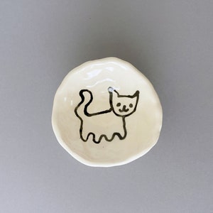 May include: White ceramic dish with a hand-drawn black cat design. The cat is facing right and has a simple, smiling face.