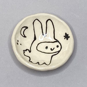 May include: A white ceramic dish with a black hand-painted design of a bunny with a crescent moon and a star.
