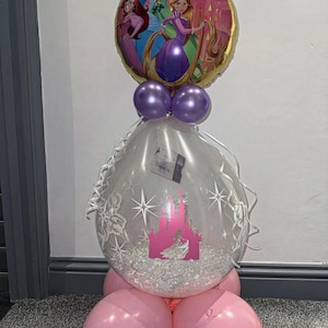 Puede incluir: Un globo transparente con un diseño de castillo rosa y purpurina en su interior. El globo está unido a cuatro globos rosas y un globo morado con un globo de lámina dorada con princesas de Disney en la parte superior.