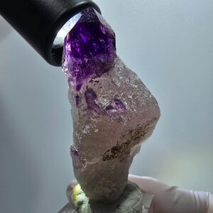 以下が含まれることがあります： 深い紫色の半透明な先端を持つ未加工のアメジスト結晶標本。先端から下部にかけては淡く濁った質感のベースへと変化しています。黒い懐中電灯が上部を照らし、結晶の自然な構造を際立たせています。