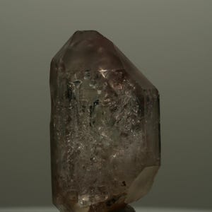 Pode incluir: Um cristal de quartzo fumê translúcido com uma terminação pontiaguda. O espécime mineral apresenta um tom castanho-acinzentado, uma estrutura geométrica facetada e um interior altamente texturizado e fraturado com inclusões internas visíveis.