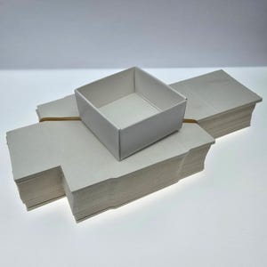 Peut inclure: Une pile de bases de boîtes en carton blanc, plates et empilées, maintenues par un élastique marron, avec une base de boîte carrée assemblée posée sur le dessus, présentant une finition mate et une construction simple et épurée.