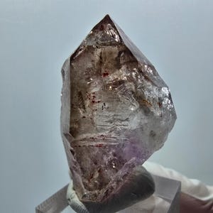 May include: A pointed, translucent quartz crystal featuring internal inclusions of reddish-brown mineral deposits and subtle purple hues at the base. The crystal has a multifaceted, jagged surface with natural fractures and clear, glassy sections.