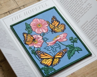 May include: A blue embroidered patch with a green border featuring three monarch butterflies and pink flowers. The text "The Hopeful" is printed above the patch and "Story" is printed below the patch. The text "Monarch Butterfly" is printed on the right side of the patch and the text "Anemone" is printed below the patch.