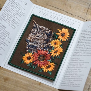 May include: An embroidered patch depicting a wolf's head surrounded by sunflowers. The patch is brown with green edging and the text "The Kindred" is printed above the image. The text "WOLF" and "SUNFLOWER" are printed below the image with descriptions of the animals and flowers.