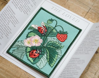 Puede incluir: Un parche bordado verde con una planta de fresa, dos mariquitas y una flor blanca. El parche está sobre un fondo azul claro con el texto "THE LOVED" en blanco.