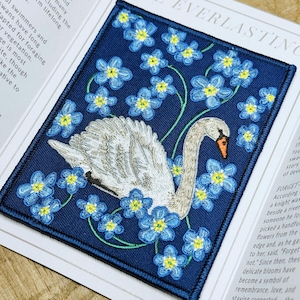 May include: Embroidered patch with a white swan and blue and yellow flowers on a navy blue background. The rectangular patch has a black border. The text "EVERLASTING" is visible. The design is detailed and intricate.