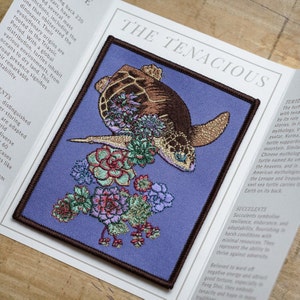 May include: Embroidered patch depicting a sea turtle with a floral design. The turtle is brown and tan, set against a blue backdrop with a brown border. The text "THE TENACIOUS" is at the top.