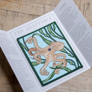 May include: A colorful embroidered patch featuring an octopus with blue eyes, surrounded by green kelp on a light blue background. The patch is square and has a green border. The text "The Adaptable" is printed on the card behind the patch.
