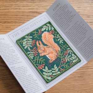 Könnte beinhalten: Gestickter Patch mit einem roten Eichhörnchen, das auf einem kleinen Holzhocker sitzt, umgeben von grünem Laub und roten Beeren. Der Patch hat einen grünen Hintergrund und den Text "THE MISCHIEVOUS" in weißen Buchstaben.
