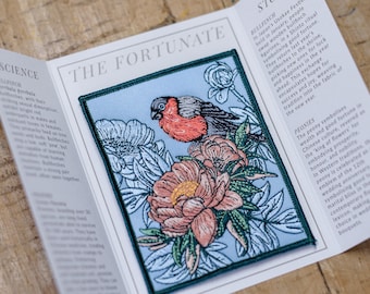 May include: A blue embroidered patch with a red-breasted bullfinch perched on a pink peony flower. The patch is surrounded by green leaves and other flowers. The text "The Fortunate" is printed on the card behind the patch.