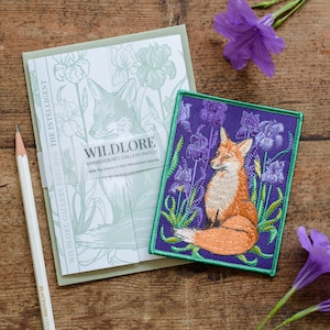 May include: A purple embroidered patch featuring a fox sitting amongst purple irises. The patch is edged with a green border. A white label reads "WILDLORE EMBROIDERED GALLERY PATCH".