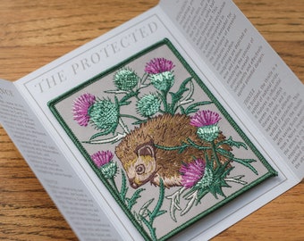 May include: A beige embroidered patch with a hedgehog curled up among purple and green thistles. The text "THE PROTECTED" is at the top of the patch. The text "STORY" is on the right side of the patch. The text "THISTLE" is at the bottom of the patch.